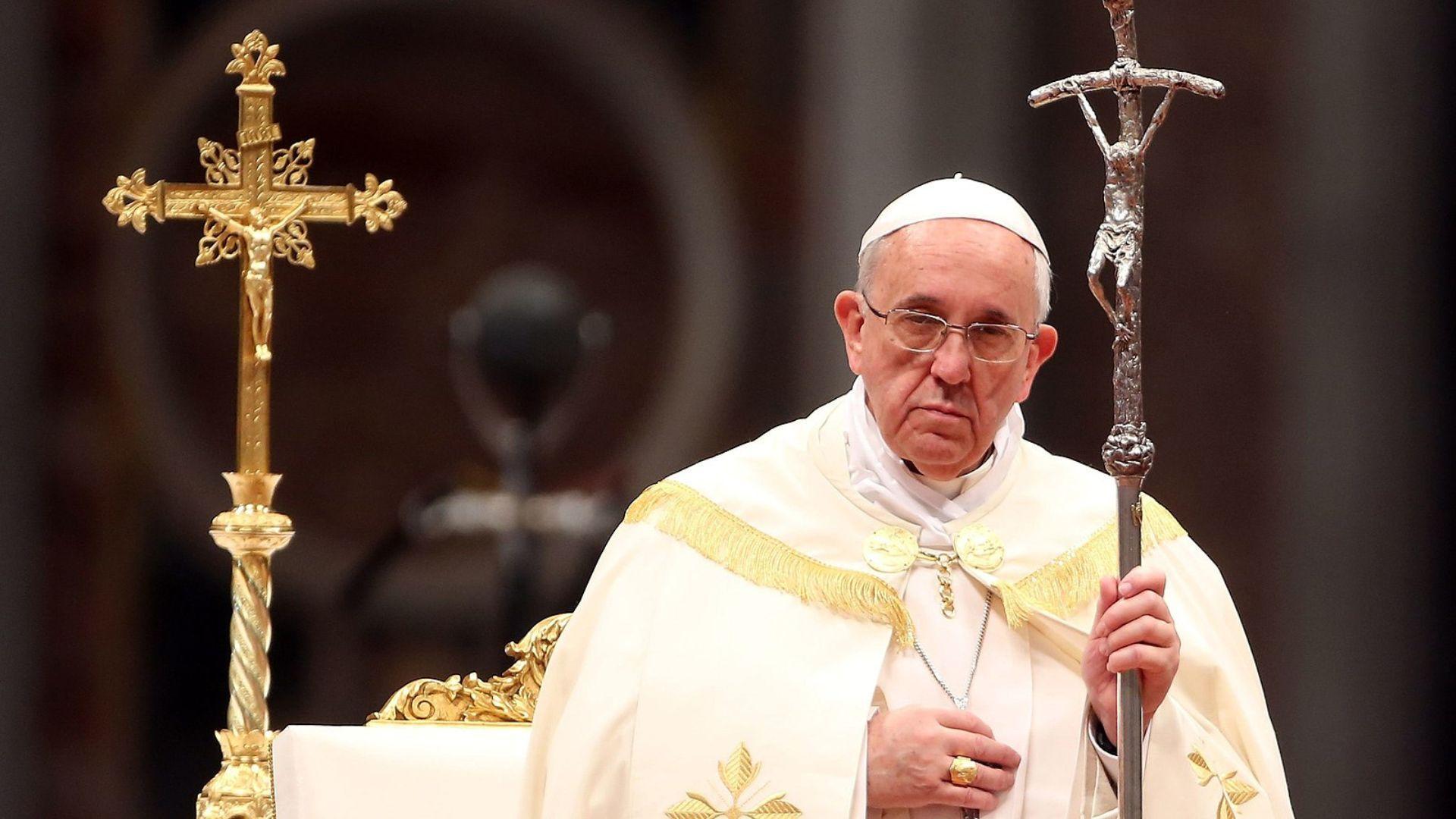 Pope Francis, wearing white papal garments with gold trim, is seen solemnly holding a staff with a crucifix on top