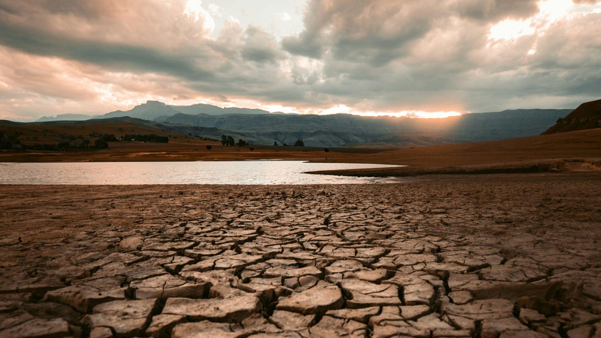 A parched, cracked lakebed in the foreground leads to a small body of water under a dramatic sunset sky