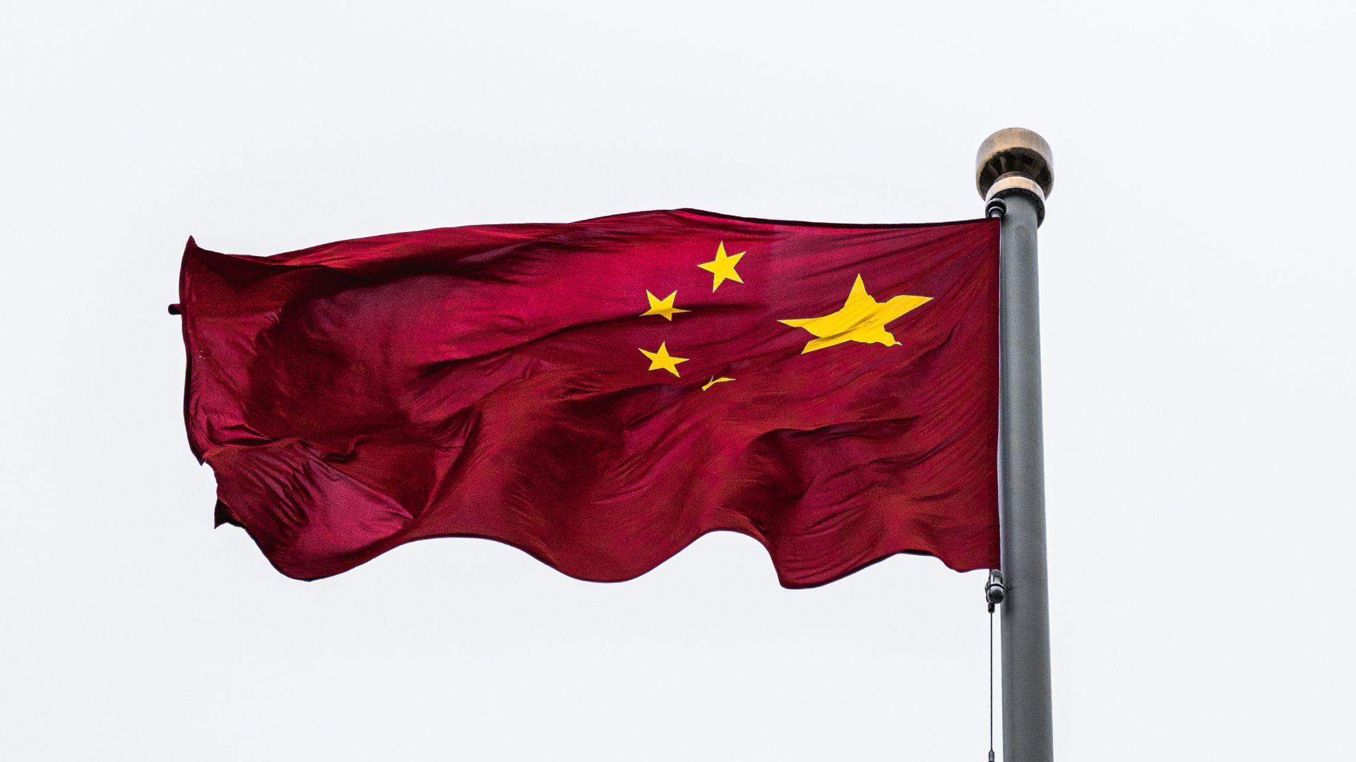 The national flag of China is unfurled and waving in the breeze, attached to a flagpole against a clear, pale sky