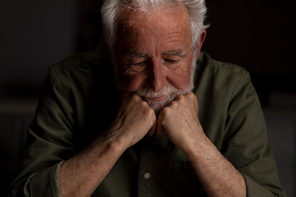An older man dressed in a green shirt who is visibly upset sits with his head in his hands