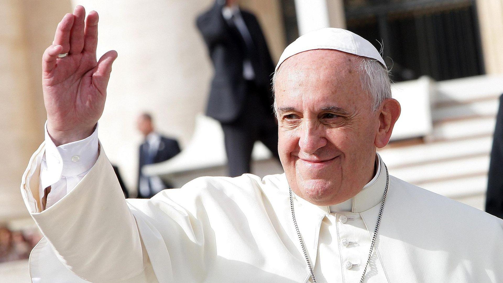 Pope Francis, dressed in his white papal attire, is captured in a candid moment, smiling and waving his hand to a crowd not visible in the frame