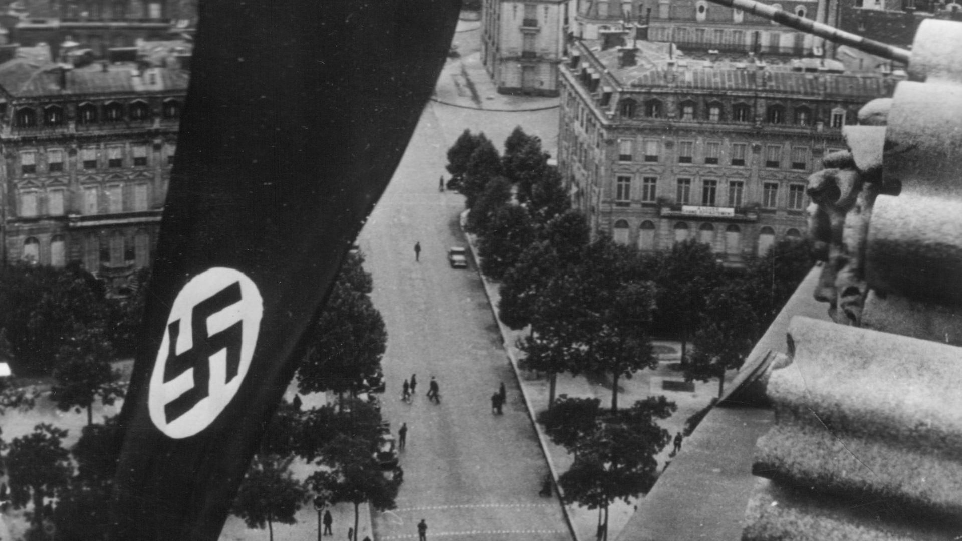 A black and white photo of a Nazi flag hanging over the streets of Paris with people walking below.