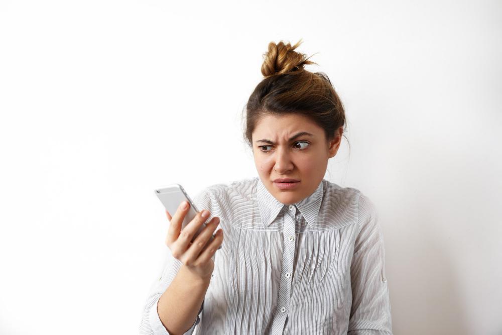 A woman dressed in a shirt looks at her smartphone with a concerned face