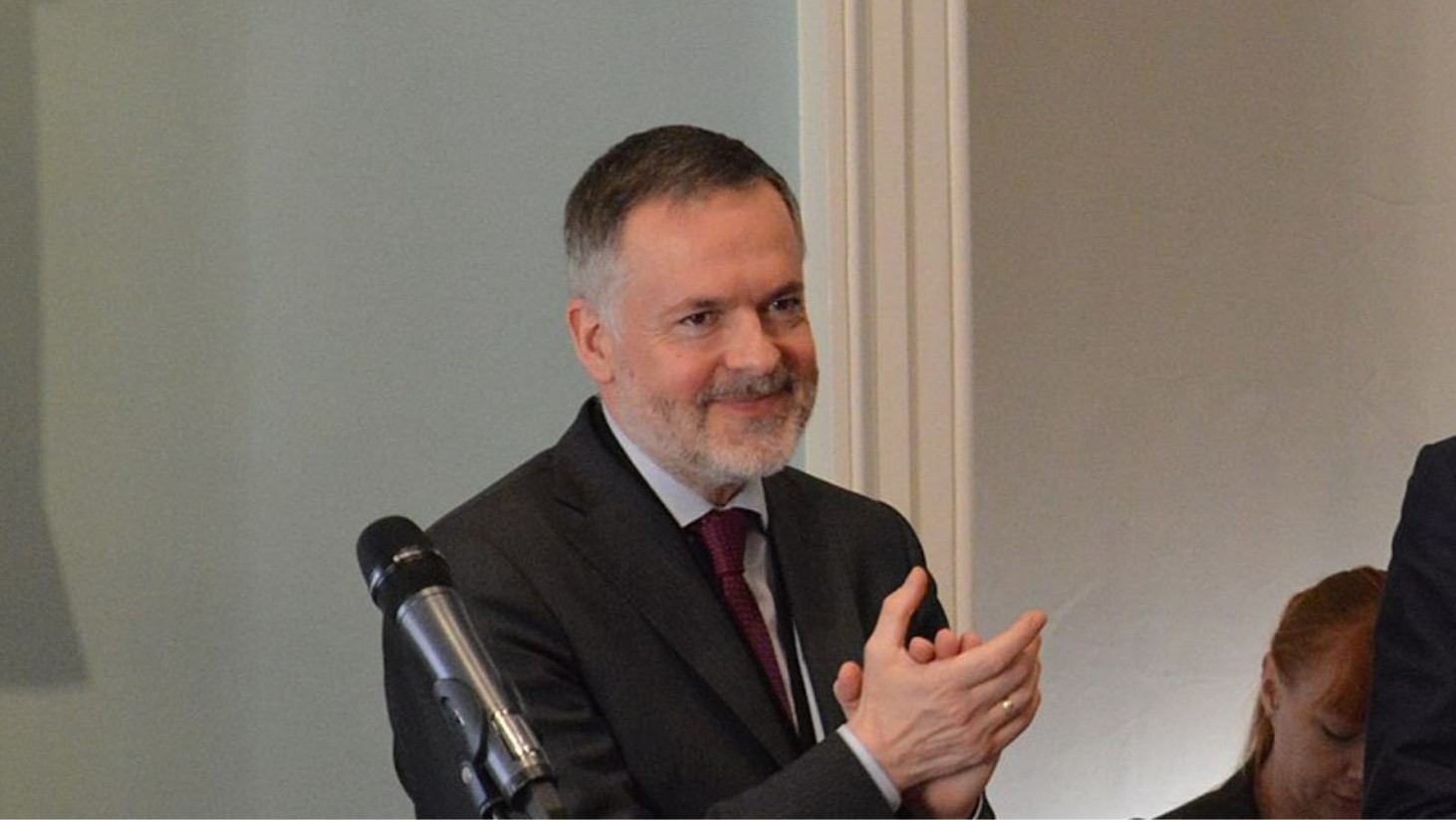 Hartwig Fischer is photographed in a dark suit, standing next to a microphone, he has his hands in a clapping position and is smiling slightly