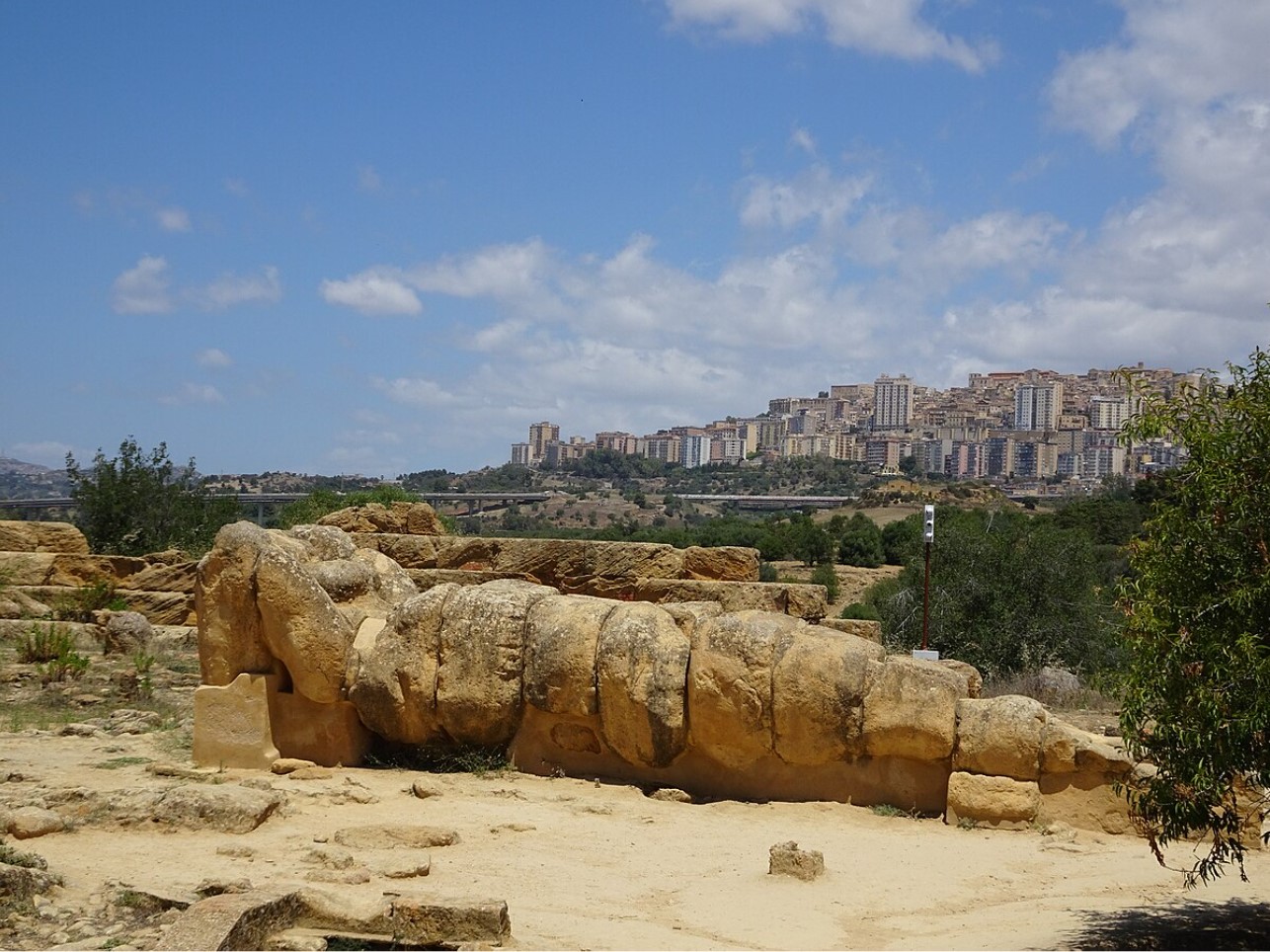 The ruins of a large male-shaped statue sits before a modern city in the distance.