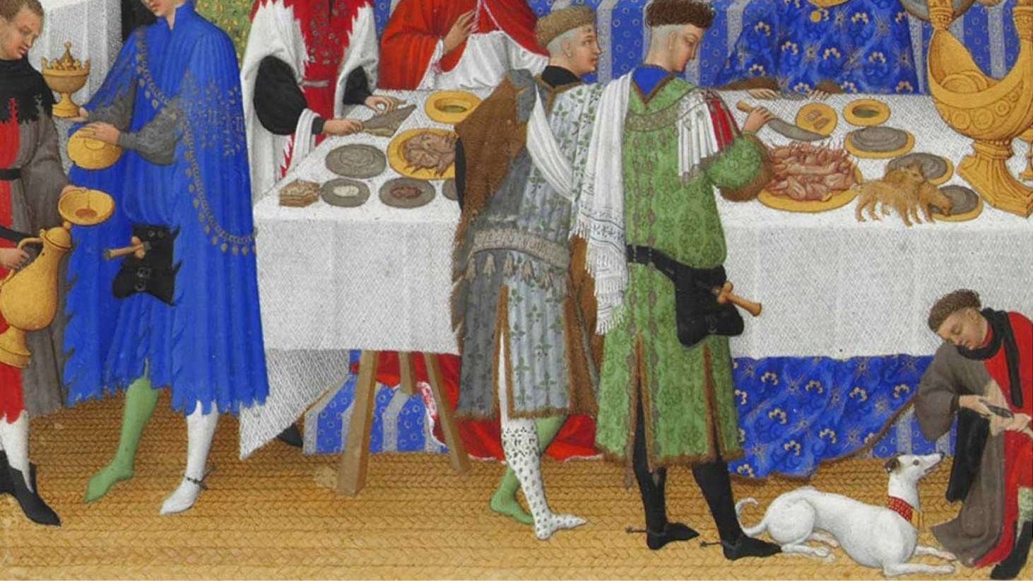 This is an illustration from a medieval manuscript depicting a banquet scene. Elegantly dressed individuals are gathered around a table covered with a white cloth, laden with various dishes and a large golden goblet. In the foreground, a person in red pours liquid into a bowl, while a dog interacts with a figure in red