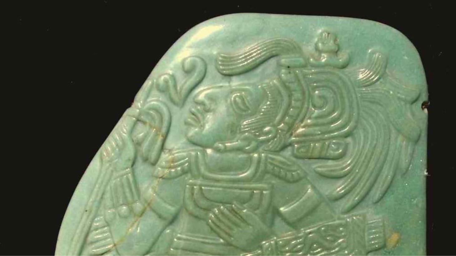 This image shows a carved jade plaque featuring Maya iconography. It displays a profile of a human figure with elaborate headdress and ear spools, indicative of high status or royalty in Maya culture
