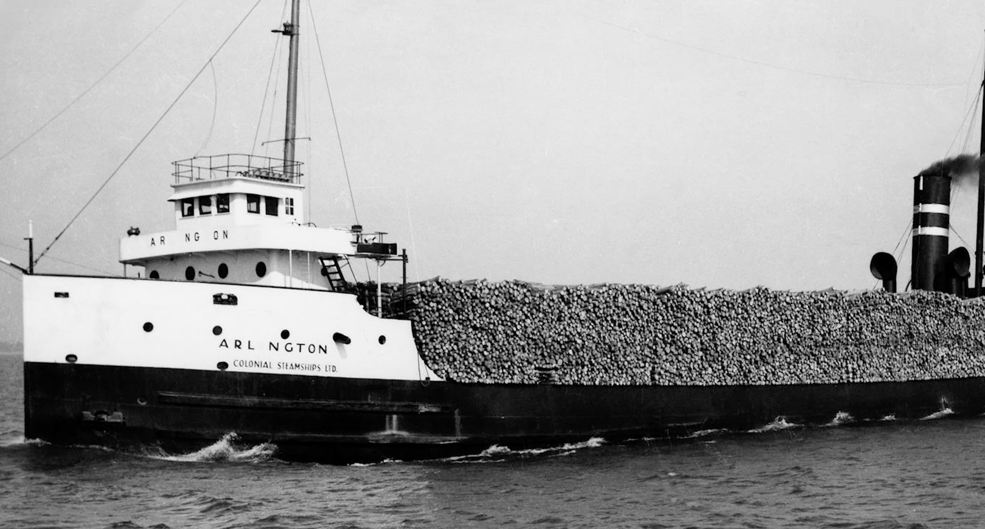 A gray scale image of a white and black ship carrying logs