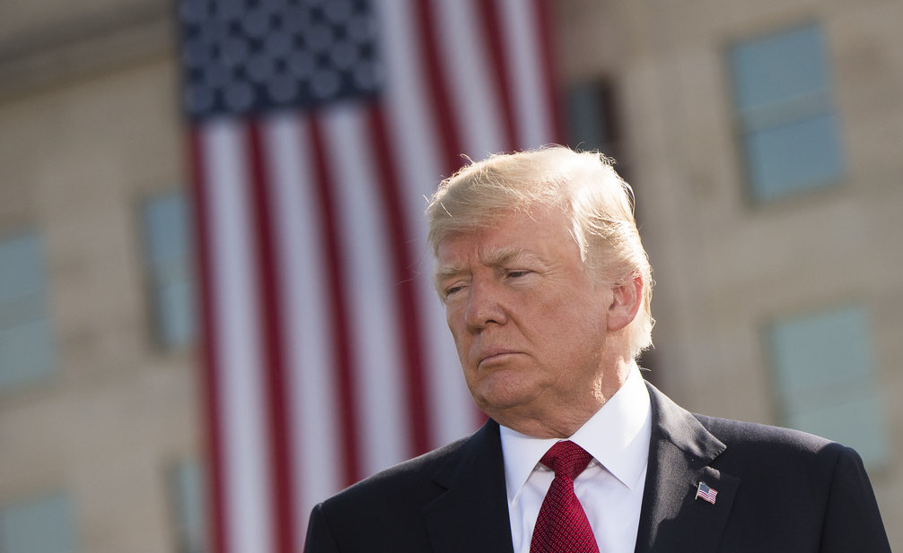 Donald Trump in a black suit with a red tie standing in front of an American flag