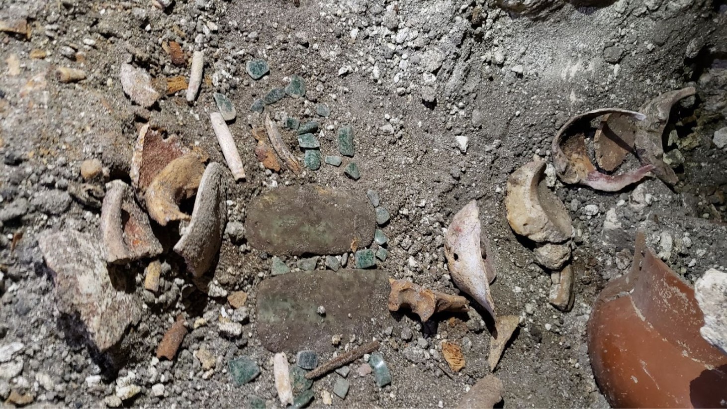 An image showing an assortment of archaeological finds scattered on the ground. The collection includes fragmented pottery shards, bits of jade, and bone fragments embedded in the soil