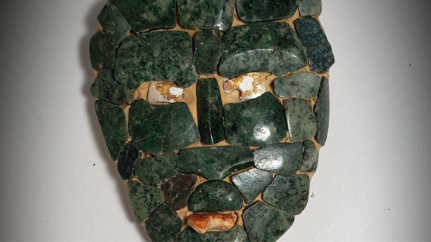 A close-up of an ancient mosaic mask made of interlocking jade pieces, with varying shades of green and some patches of beige where the jade has fractured or eroded, set against a white background