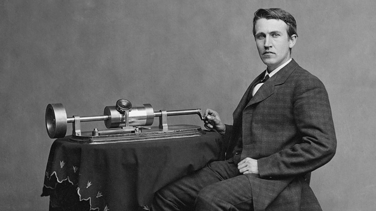 A black and white photograph of Thomas Edison sitting beside his invention, the phonograph. Edison is captured in a profile view, looking to the right, and dressed in a dark three-piece suit with a bow tie