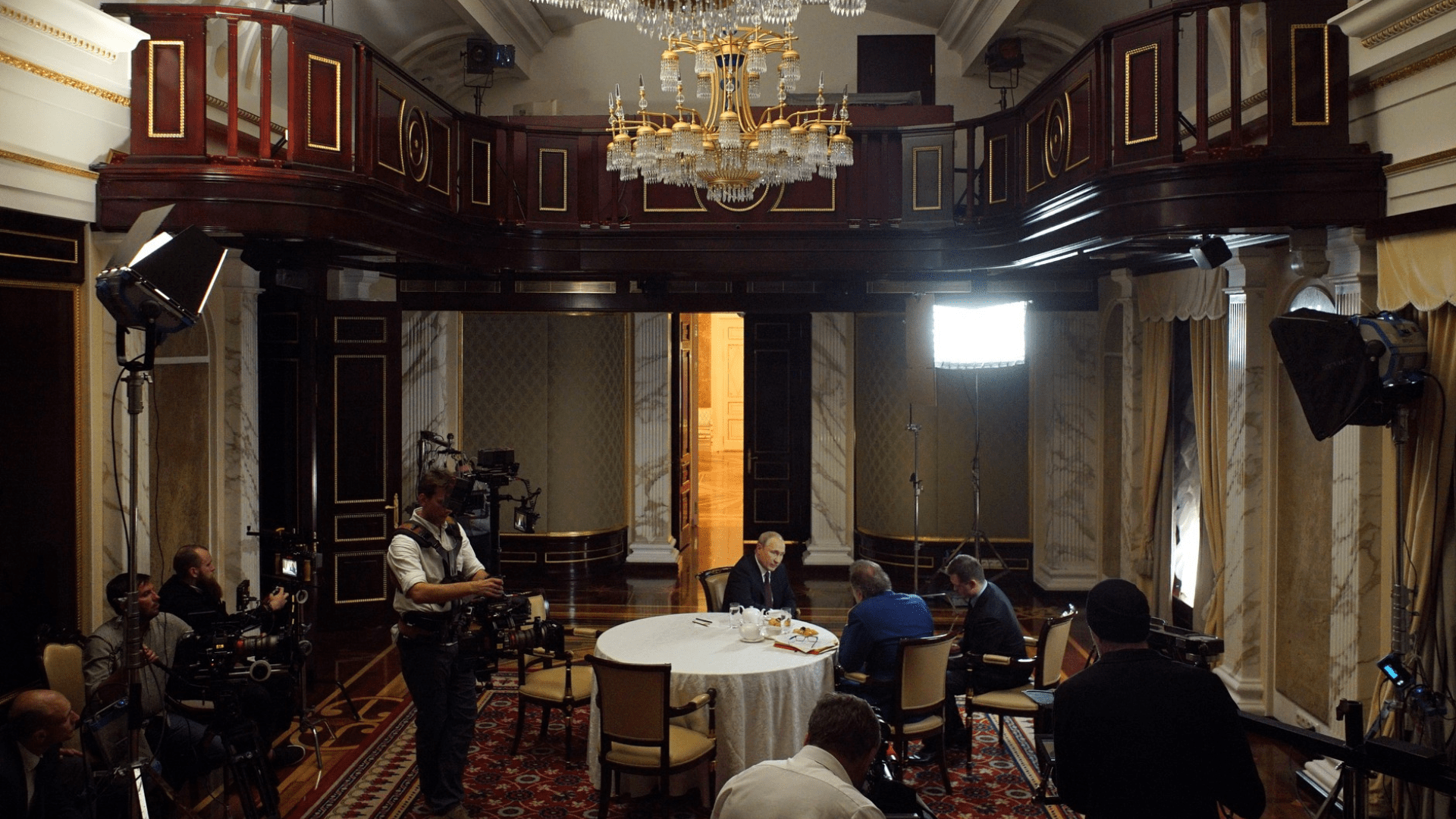 Putin sits down with Oliver Stone for an interview inside of a luxurious dining hall. Camera operators and lighting equipment surround them.