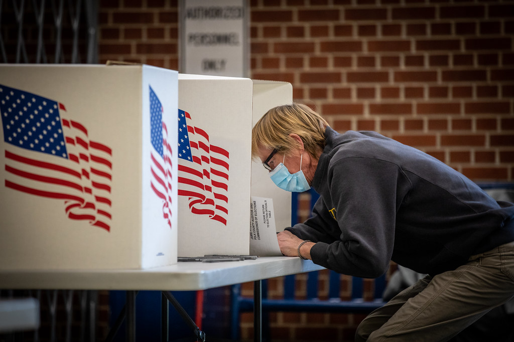 A person wearing a blue mask leaning over a table voting on a ballot