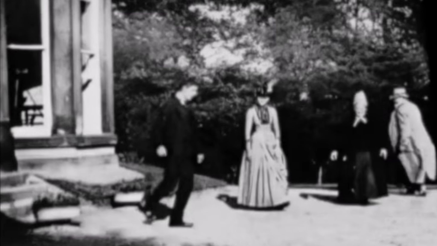 A blurry black and white still from the historic short film "Roundhay Garden Scene" depicting four individuals, two men and two women, outdoors. They are dressed in 19th-century attire; the women wear long dresses and the men in coats and trousers
