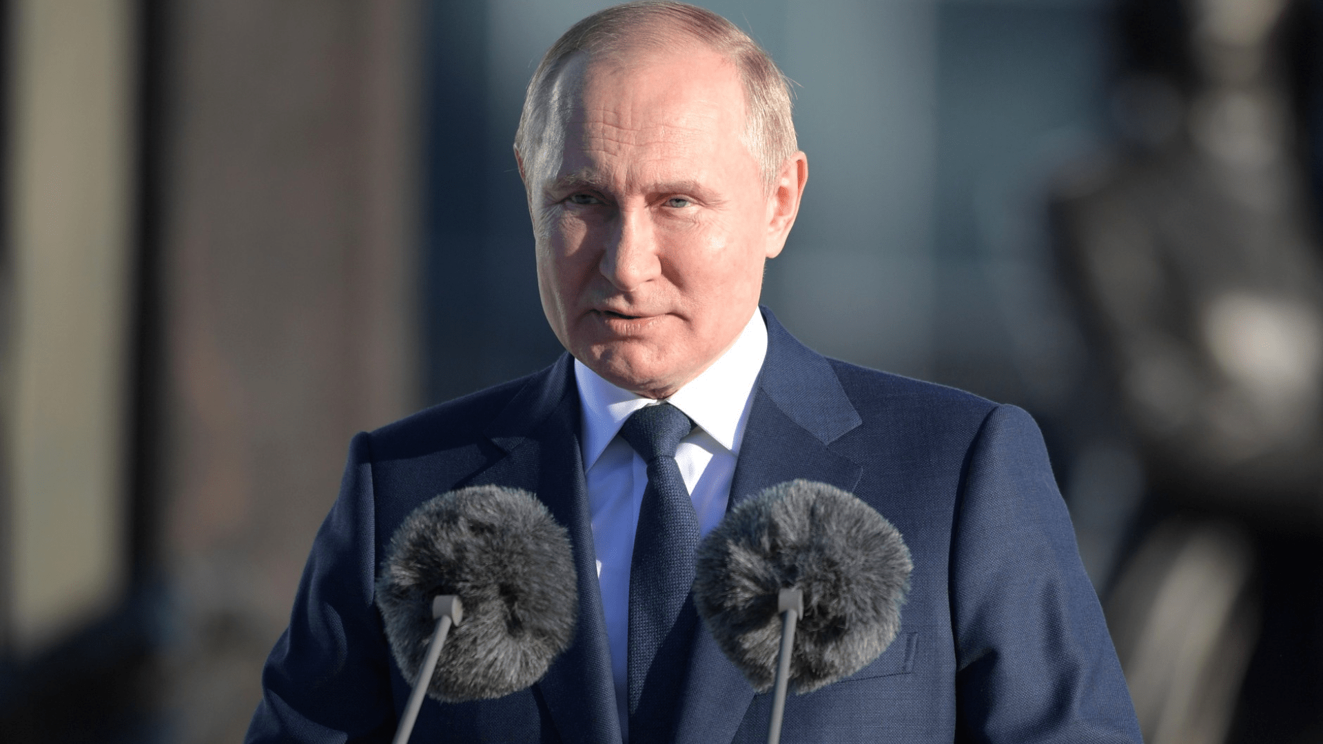 Vladimir Putin wearing a suit and speaking in front of two microphones.