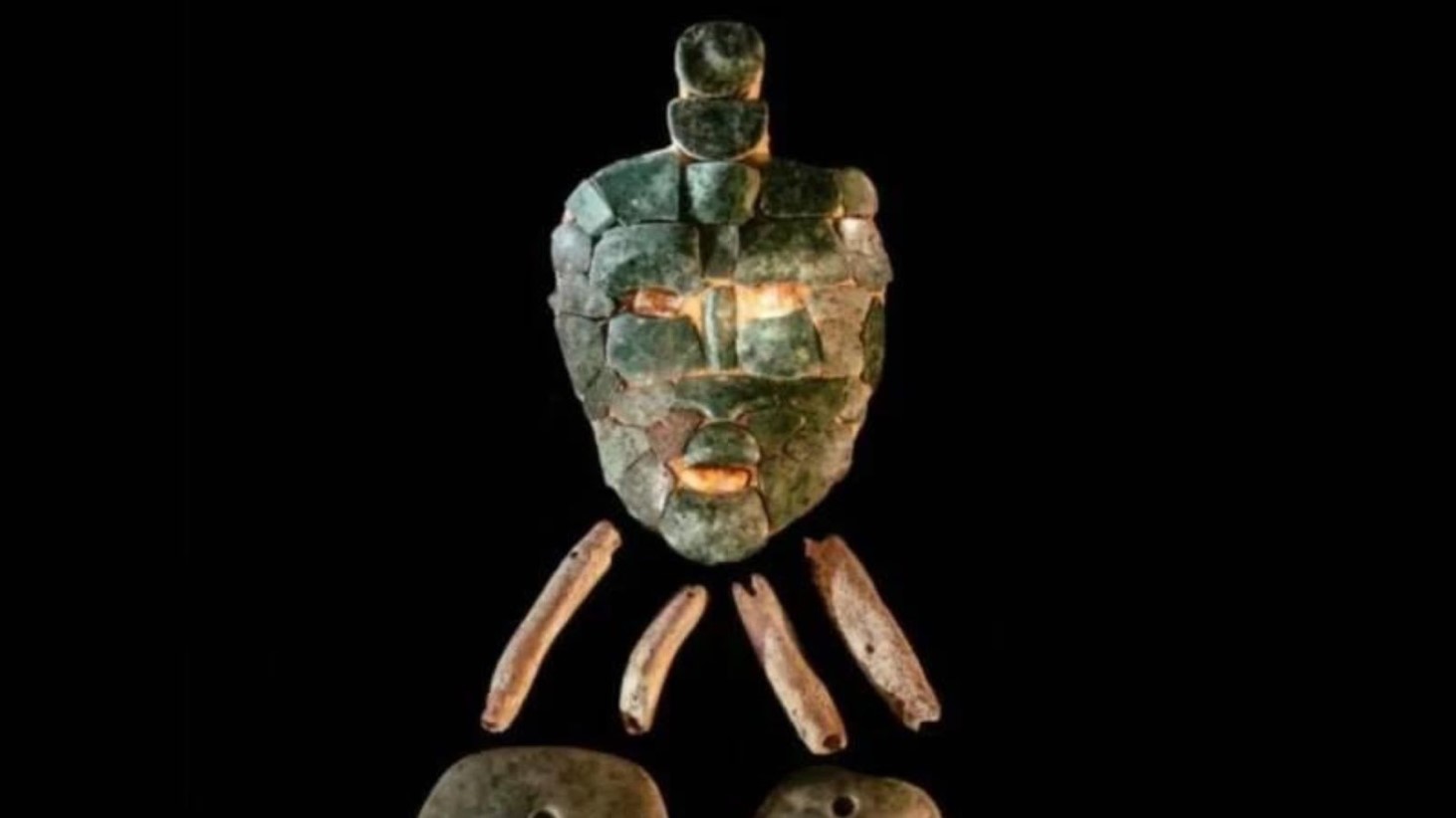 The image displays an ancient mosaic mask made of jade pieces arranged to form the facial features, including eyes, a nose, and a mouth, set against a black background. Below the mask, there are bone fragments and two round stone objects