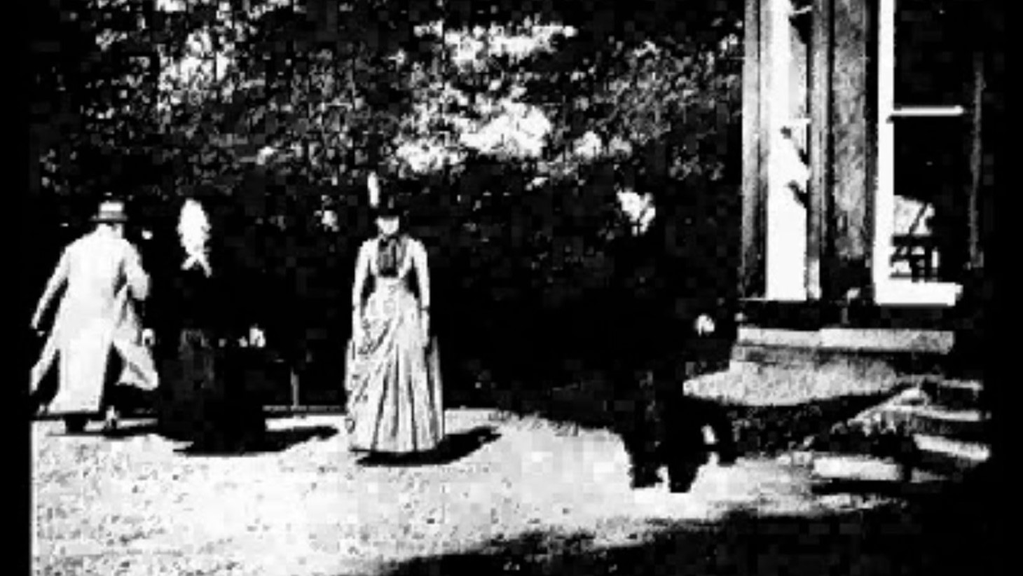 A high-contrast black and white still from the 1888 short film "Roundhay Garden Scene." It shows four figures, two men in suits and two women in full-length dresses, captured mid-movement in a garden next to a building with tall windows. The image is grainy and not sharply defined, reflecting the early technology
