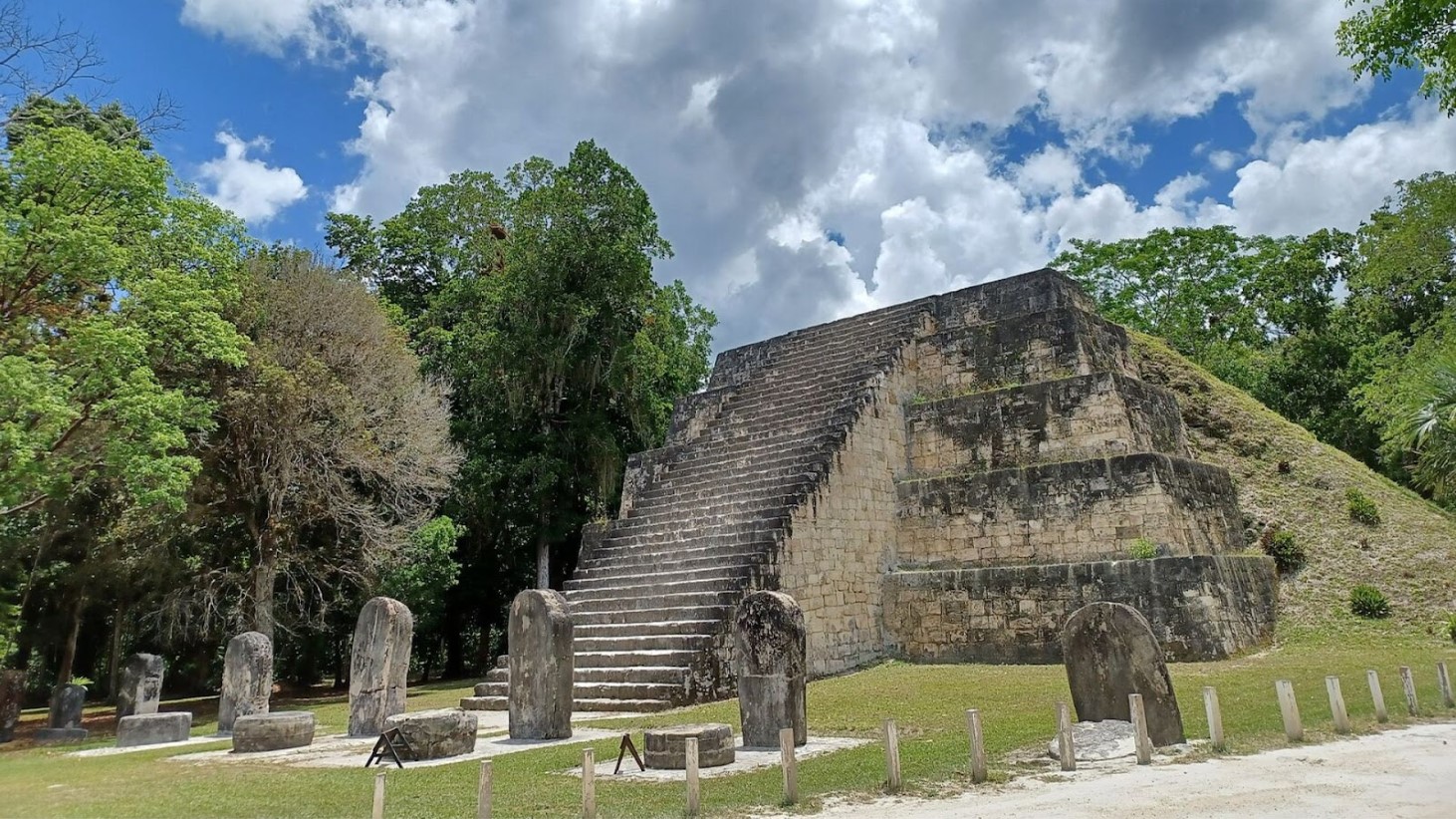 A photograph captures a historical Maya temple with a grand staircase, set against a backdrop of lush green trees and a bright blue sky with scattered clouds