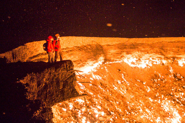 Two people stand above the Darvaza Crater on an outcropping, admiring the inferno.