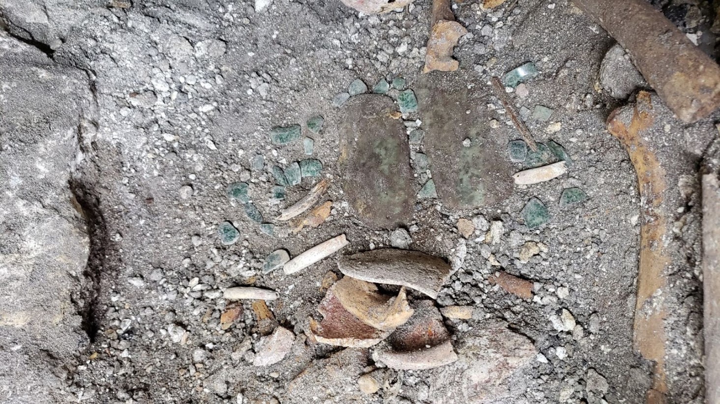 An image showing fragments of pottery, pieces of jade, and small bones embedded in a patch of grey soil. The artifacts appear scattered and partially buried