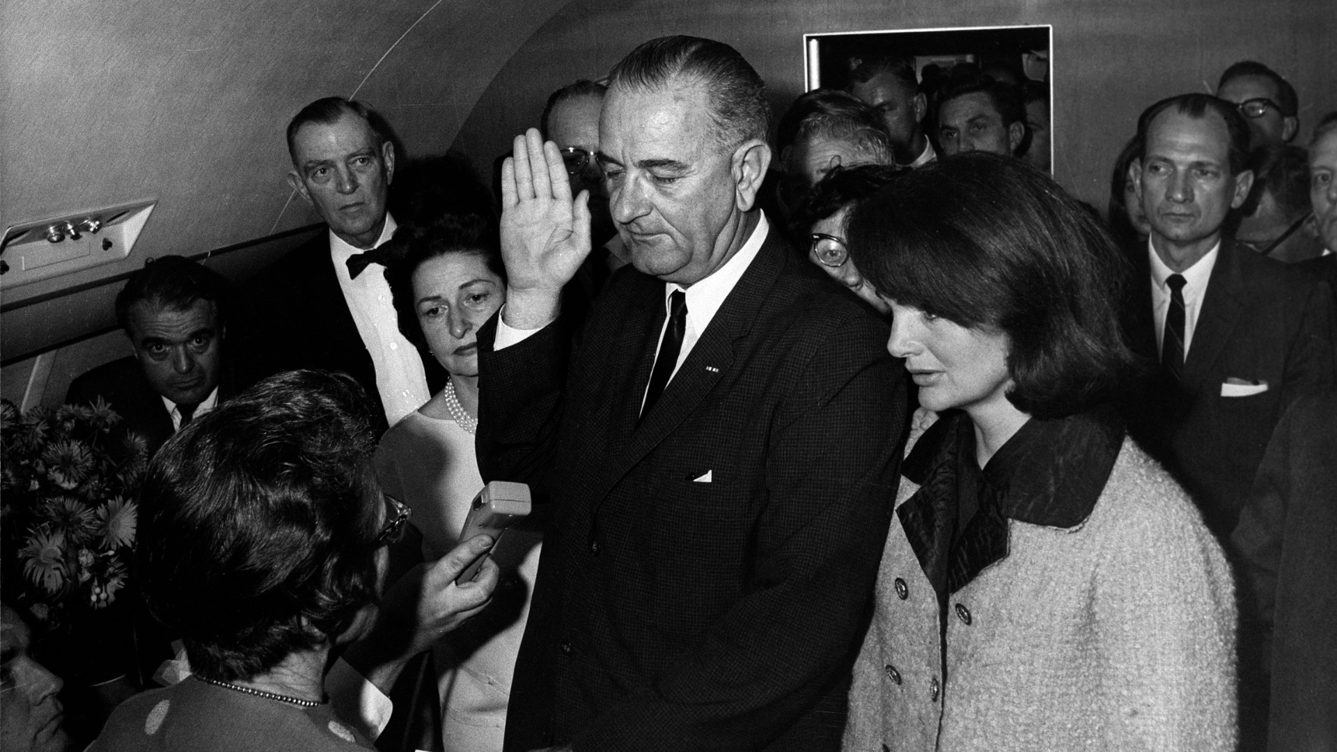 Black-and-white photo of Lyndon B. Johnson taking the oath of office aboard Air Force One two hours after John F. Kennedy’s assassination. Jackie Kennedy stands next to him.