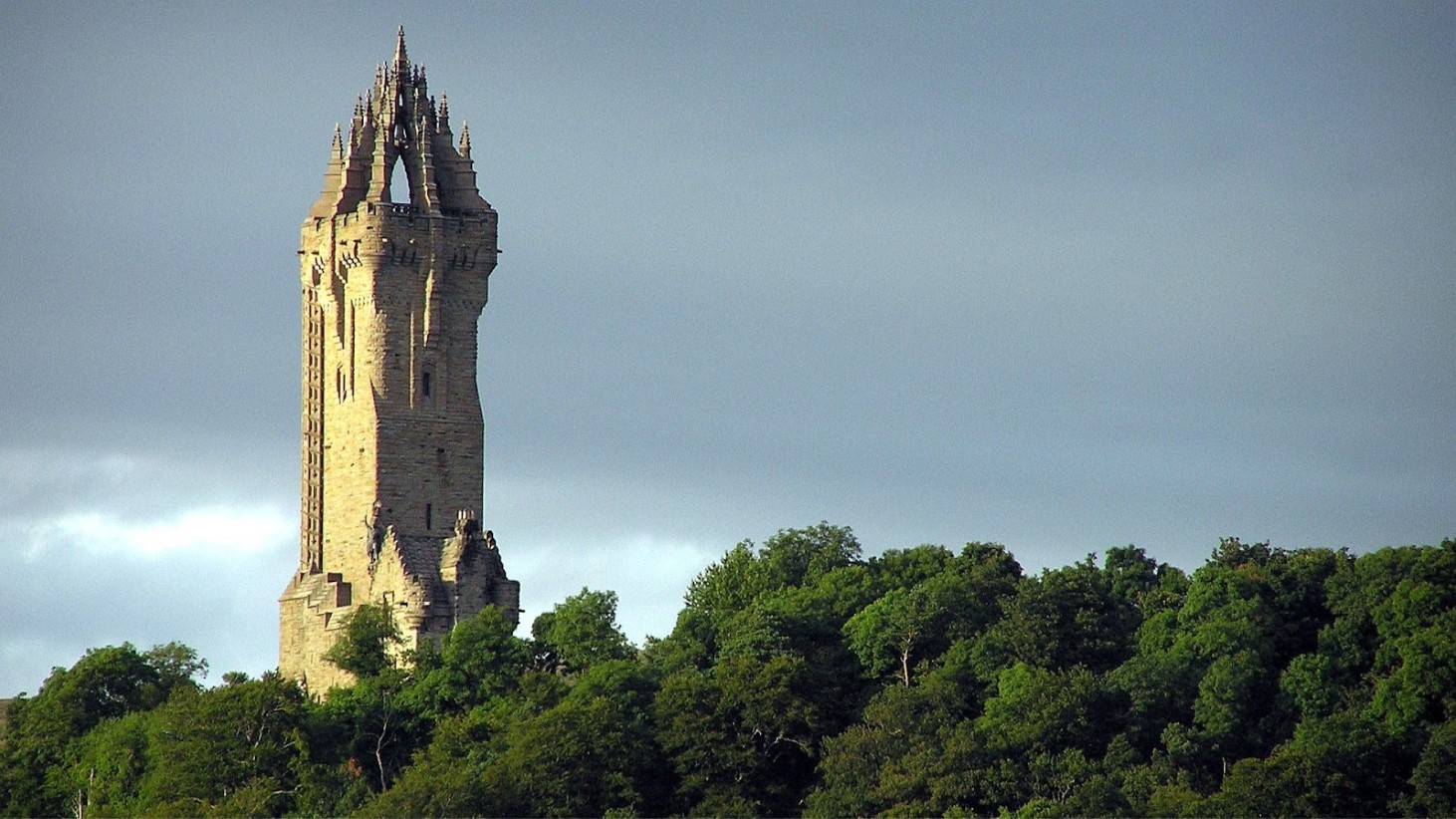 A photograph of the National Wallace Monument, a tall and slender tower rising above a lush green forest under a dramatic sky