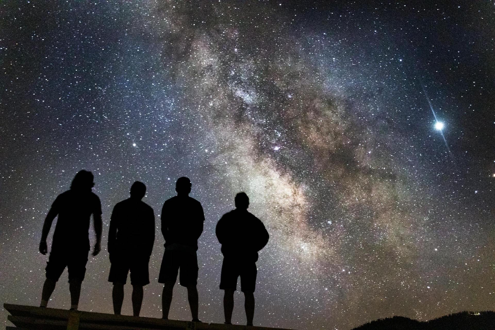 Four people standing on a hilltop looking at the Milky Way galaxy in the night sky