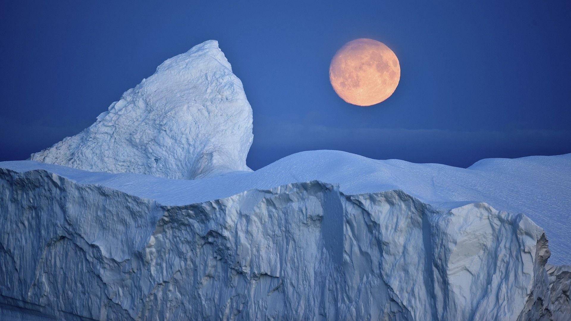 The moon rises in the sky before an iceberg located near Antarctica.