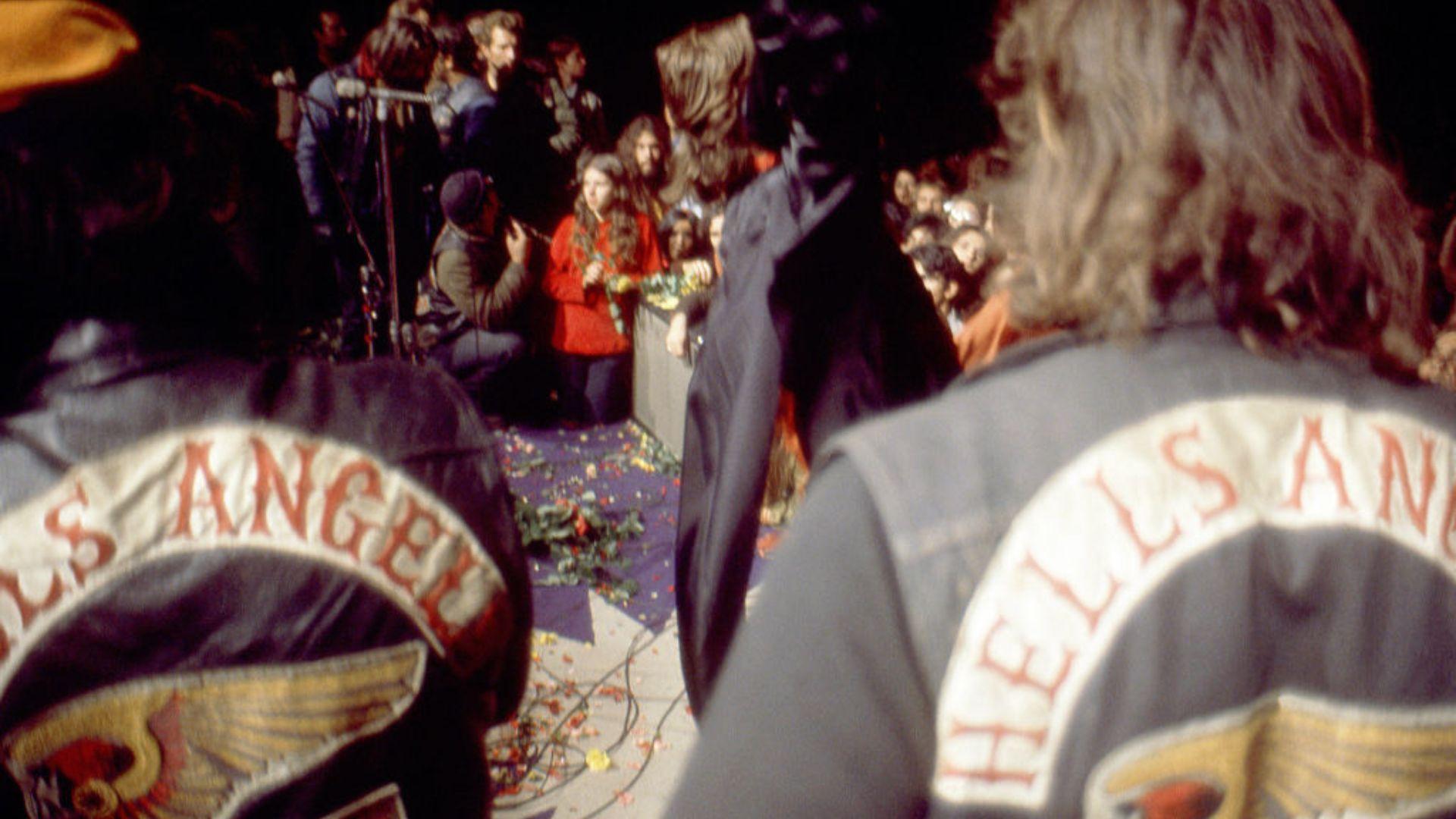 Two individuals with their backs to the camera are wearing jackets with 'Hells Angels' patches. They are facing a crowd at a concert, where people appear to be watching a performance