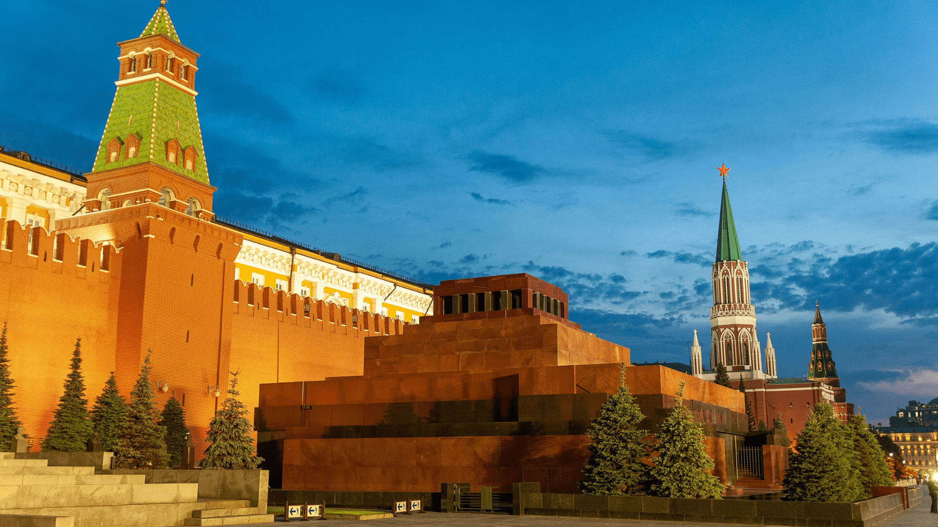 Another angle of Lenin’s tomb that shows evergreen trees growing nearby and more of the Kremlin. The walls of the Kremlin are lit up and it is dusk.
