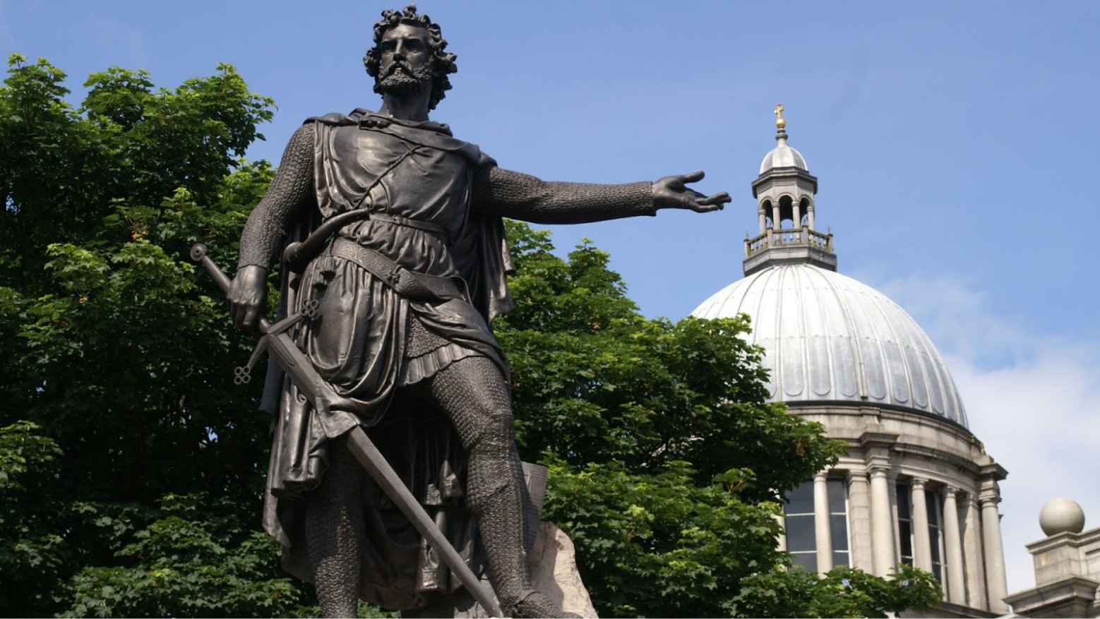 A dark William Wallace statue by green trees and an old building in Aberdeen.