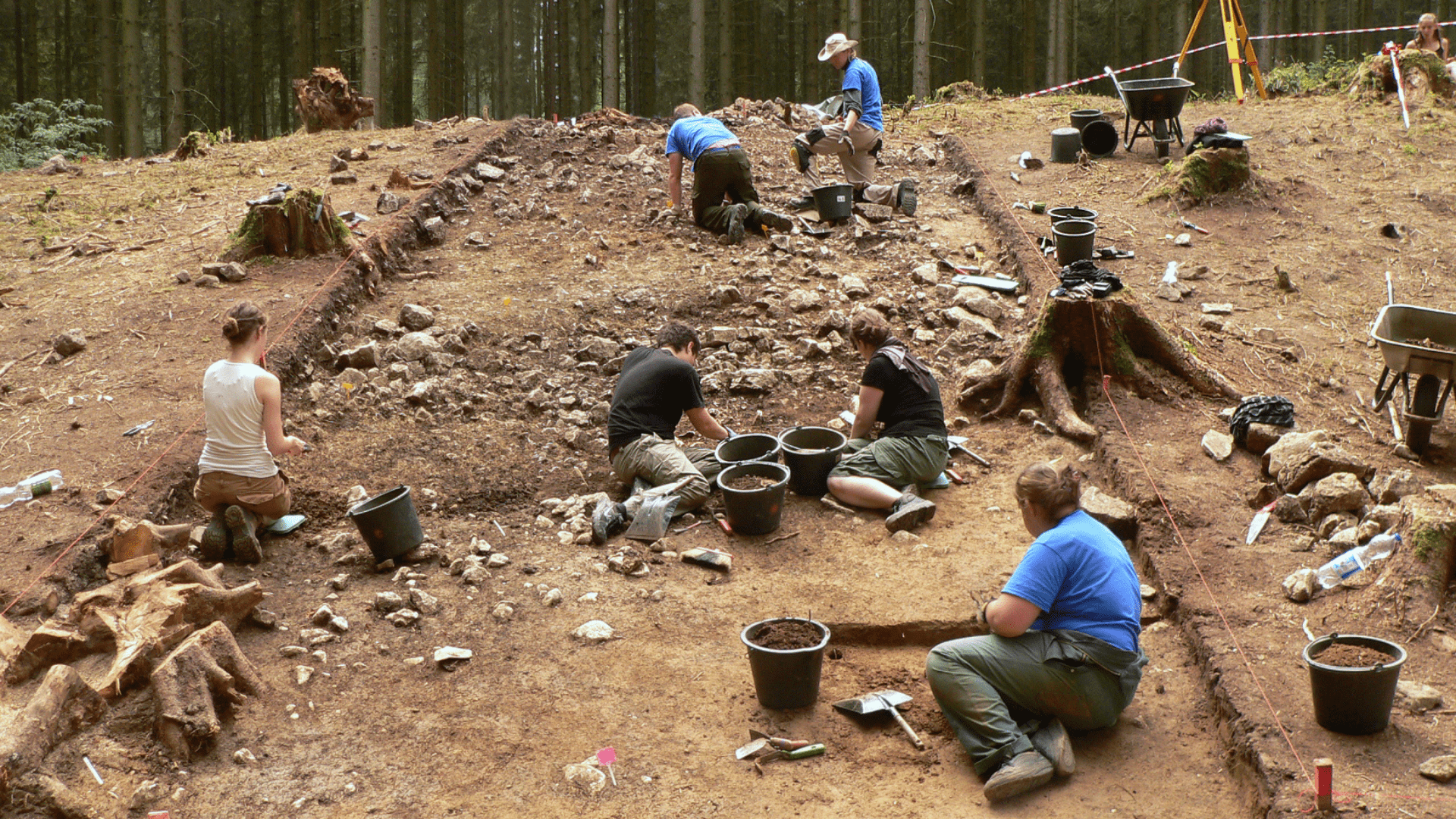 People working together on a hillside during an archaeological dig. They have special tools and buckets and are carefully digging and sifting through the dirt.