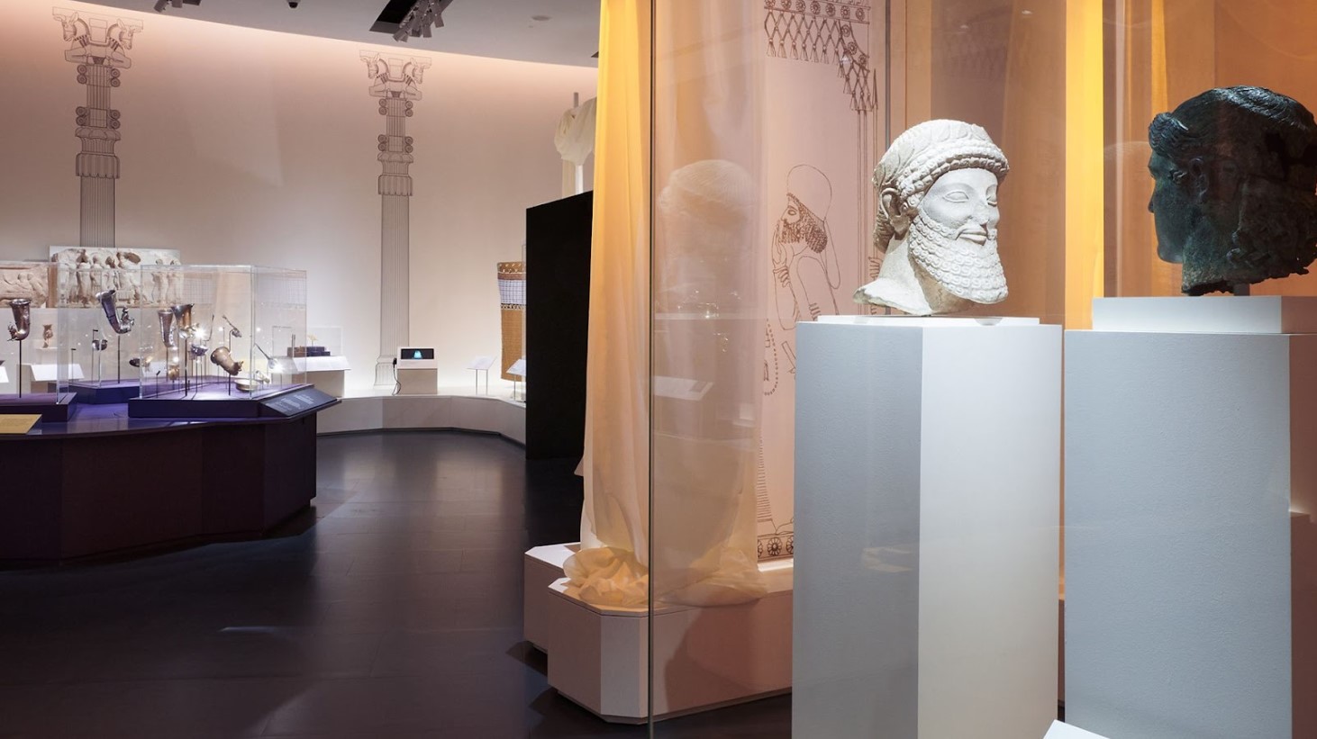 An exhibit within a museum gallery, featuring ancient artifacts, including sculptures and pottery. In the foreground, two stone heads, one of a bearded figure, are prominently displayed on pedestals. The gallery has a modern design with dark flooring and neutral walls
