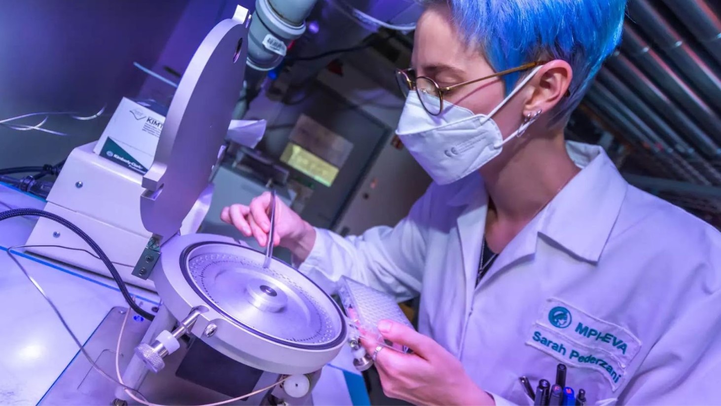 A scientist with blue hair, wearing glasses, a face mask, and a white lab coat labeled "Sarah Pederzani" is using sophisticated laboratory equipment. The scientist is carefully manipulating a sample on a machine