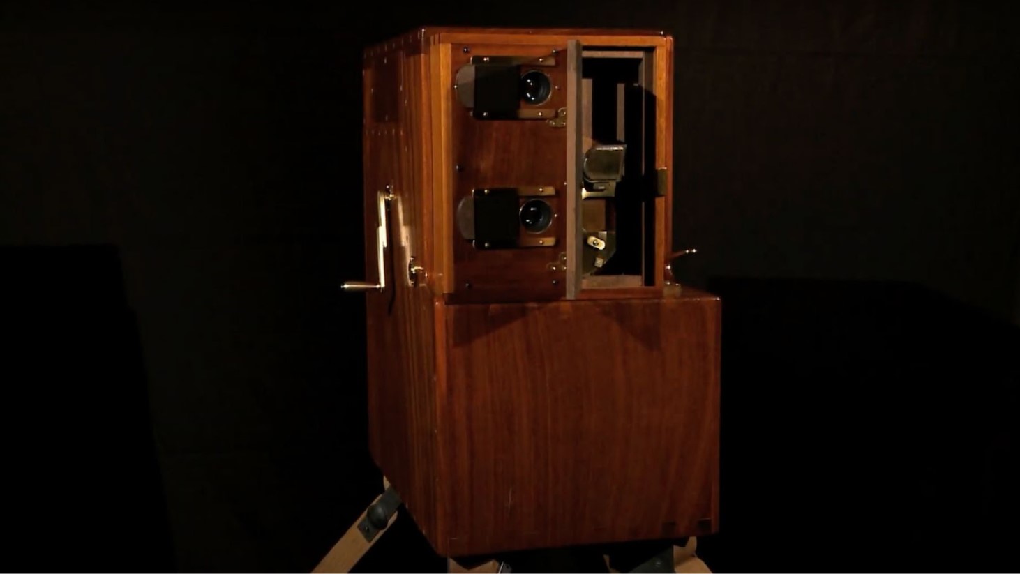 Photograph of Louis Le Prince's single-lens camera, a pioneering piece of motion picture technology from the 19th century. The camera is constructed of polished wood and features metal components, with two lenses visible on the front. It's mounted on a tripod and set against a dark backdrop