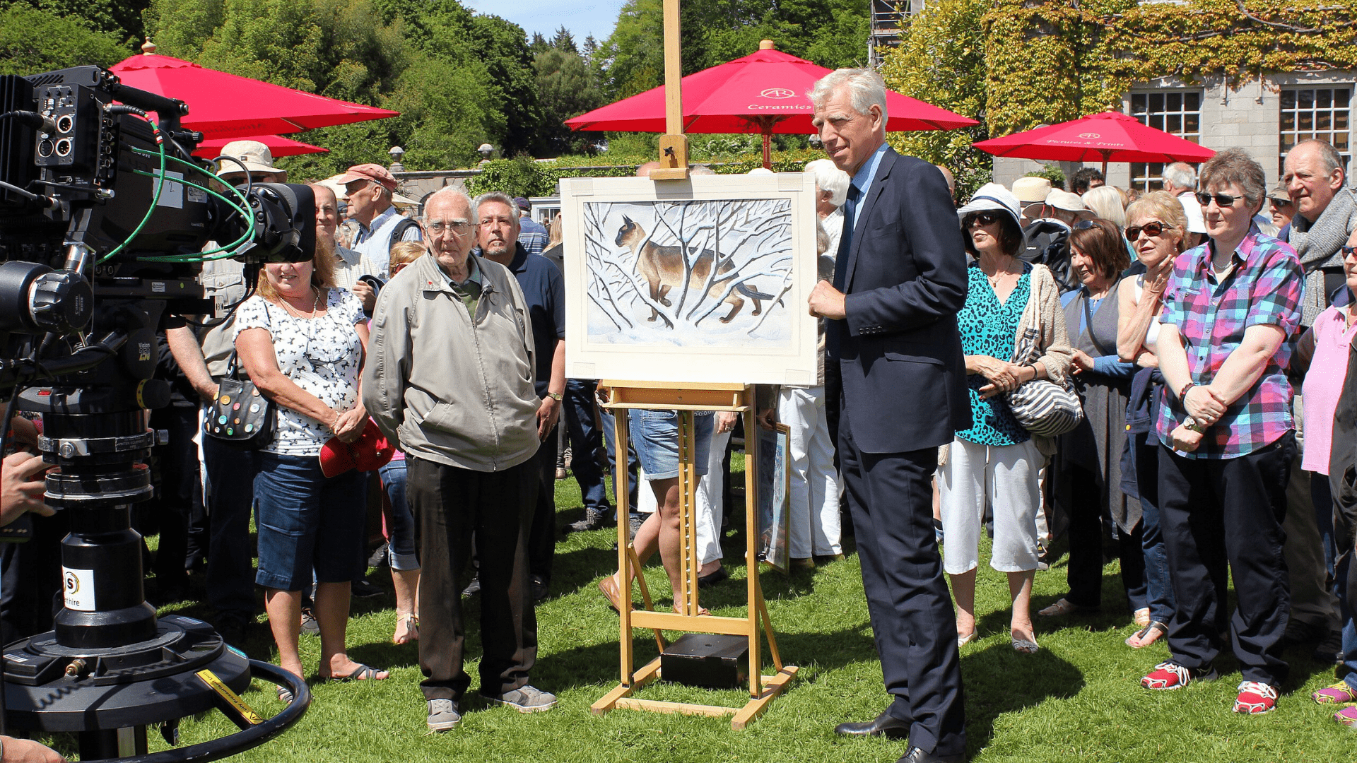 Photo from the 2015 Antiques Roadshow at Plas Newydd in Wales. A television camera is seen on the left, with an antiques expert in a suit standing next to a painting on an easel and a small crowd of onlookers standing behind the painting.