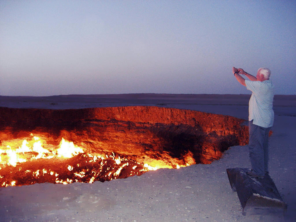 An elderly tourist takes a photo of the Darvaza Crater, raising his camera high above his head and standing on a nearby object to gain height.