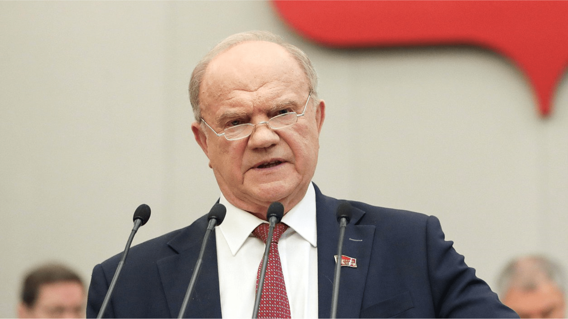 Russian Communist Party leader Gennady Zyuganov wearing a suit and speaking behind four microphones.