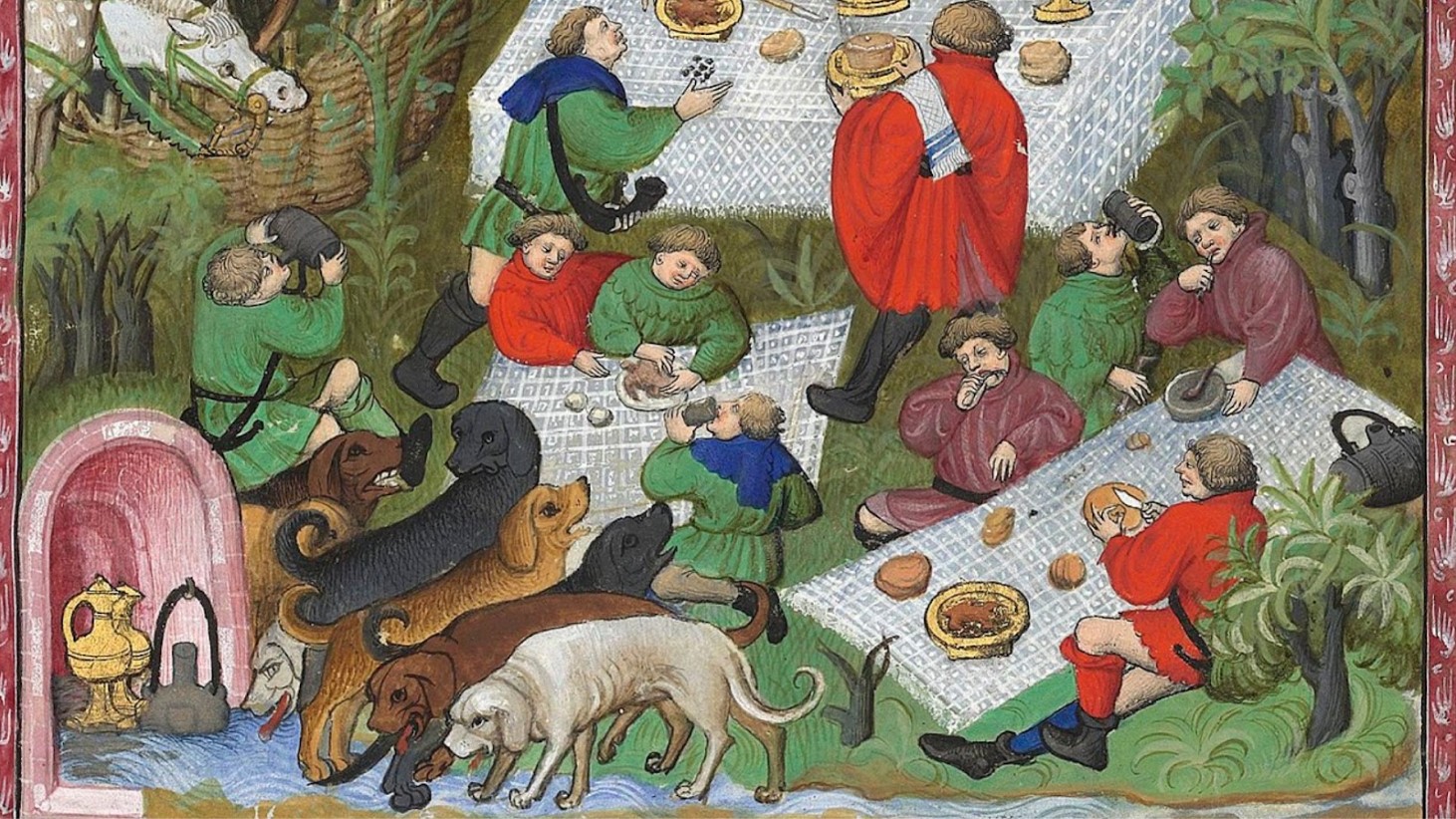 This is an image from a medieval illuminated manuscript depicting a lively outdoor feast scene. A group of individuals in colorful medieval attire are seated around a white tablecloth, enjoying various foods and drinks. Among them, dogs of different breeds are interspersed, with some drinking from a fountain in the foreground