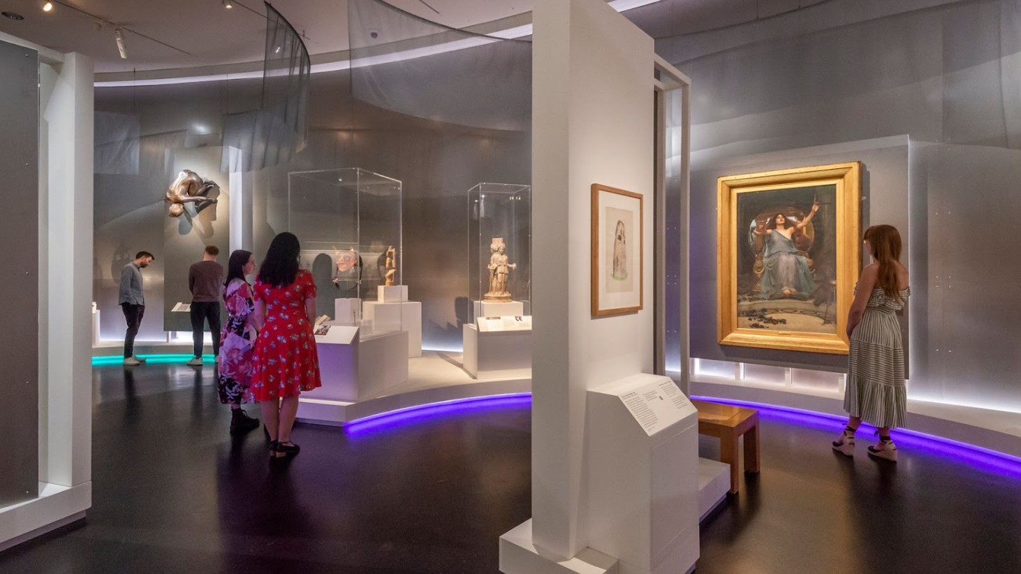 Visitors are observing various artworks in the British Museum. The room is illuminated with soft lighting and accentuated by a neon purple light along the floor. Artworks, including sculptures in glass cases and framed paintings, are displayed throughout the space