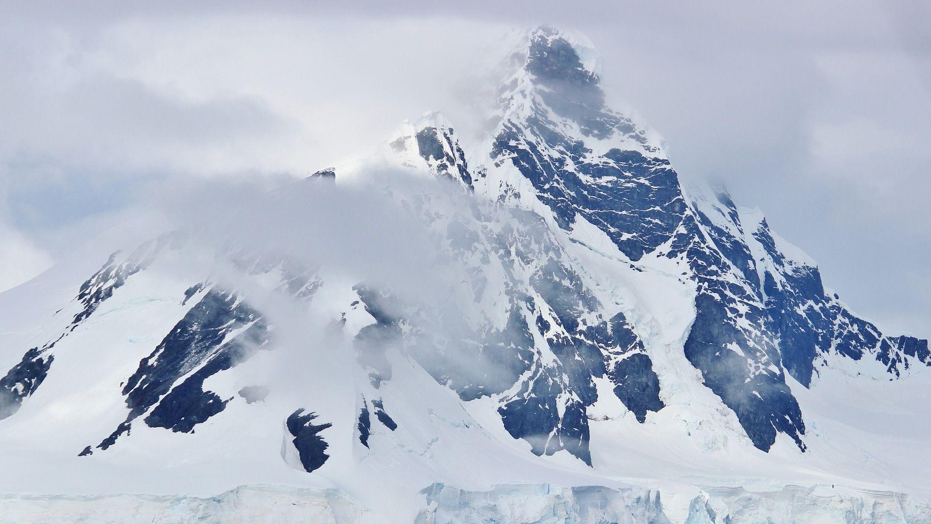 A mountain in Antarctica covered in snow and ice, slightly hidden by clouds and a hazy, grey sky.