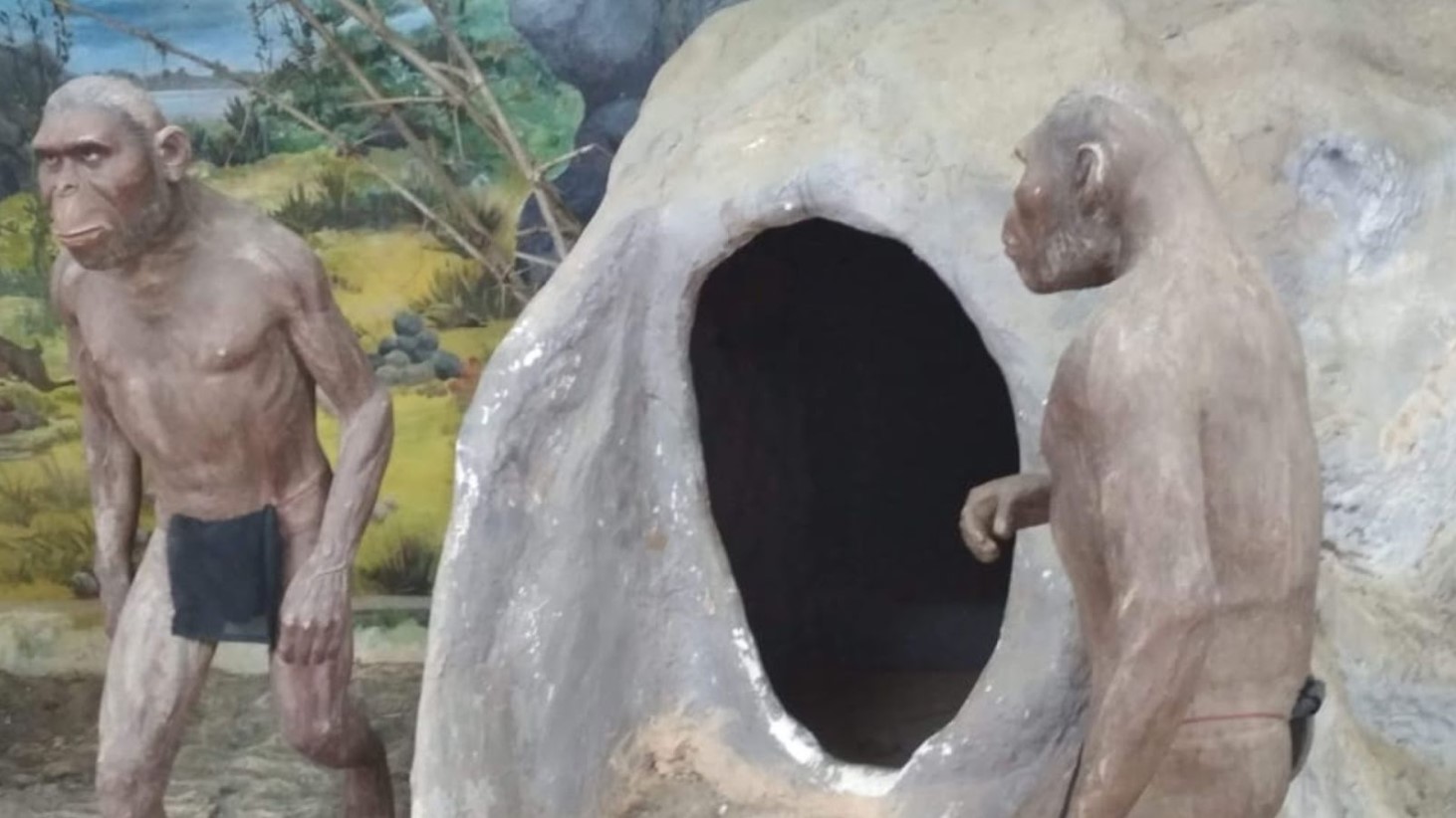 A diorama depicting two human-like ancestral figures near a rock shelter. The figure on the left stands upright, facing the viewer, with a contemplative expression and arms relaxed by its sides. The figure on the right is positioned to the side of a large, rounded opening of the shelter