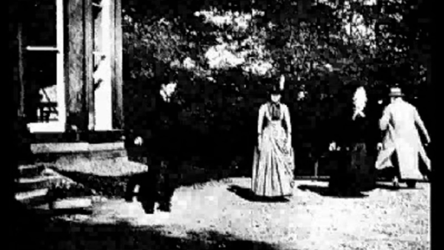 A grainy black and white screenshot depicts four individuals, two women and two men, in early 19th-century attire outside a building with large pillars. One woman stands in the foreground, centered and facing the camera, while the other woman and man appear to be walking past her