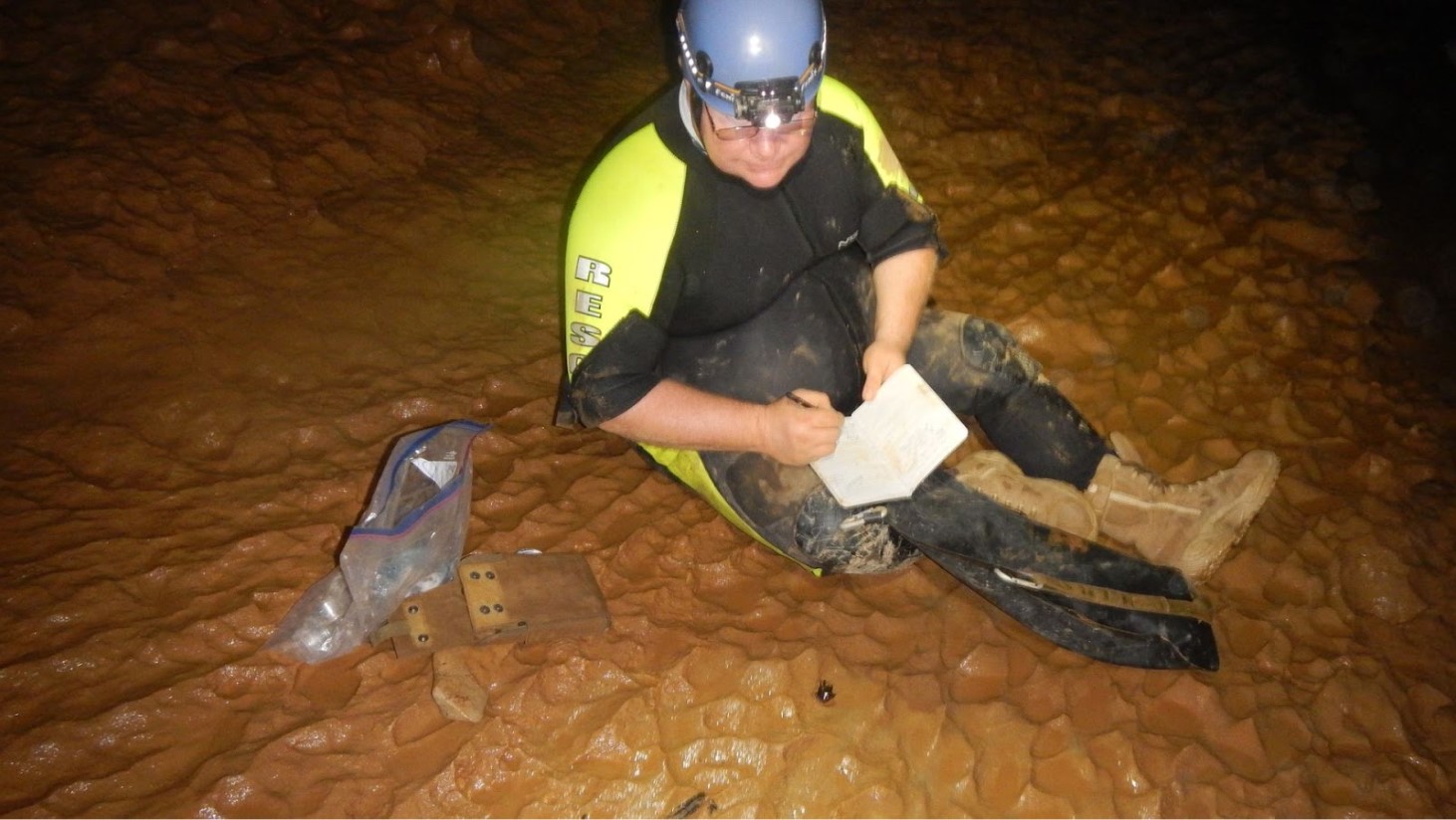 A cave researcher, wearing a wetsuit and safety helmet, is seated on the muddy floor of a cave. They are focused on writing in a notebook, with a clear plastic bag and wooden measuring tools nearby