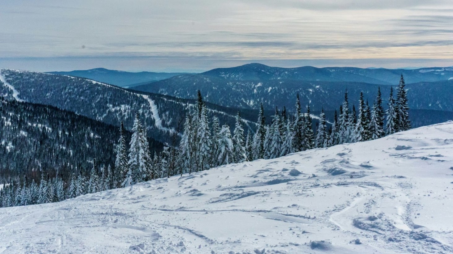A panoramic view of a snowy mountain landscape under an overcast sky. The foreground shows a slope covered in snow