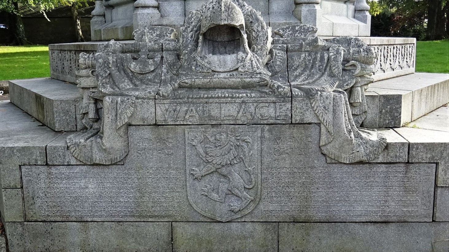 The photograph shows a close-up of the base of a monument dedicated to William Wallace, with the name 'WALLACE' prominently inscribed in the stone. Centered below is a sculpted Scottish lion rampant crest, etched into the stone