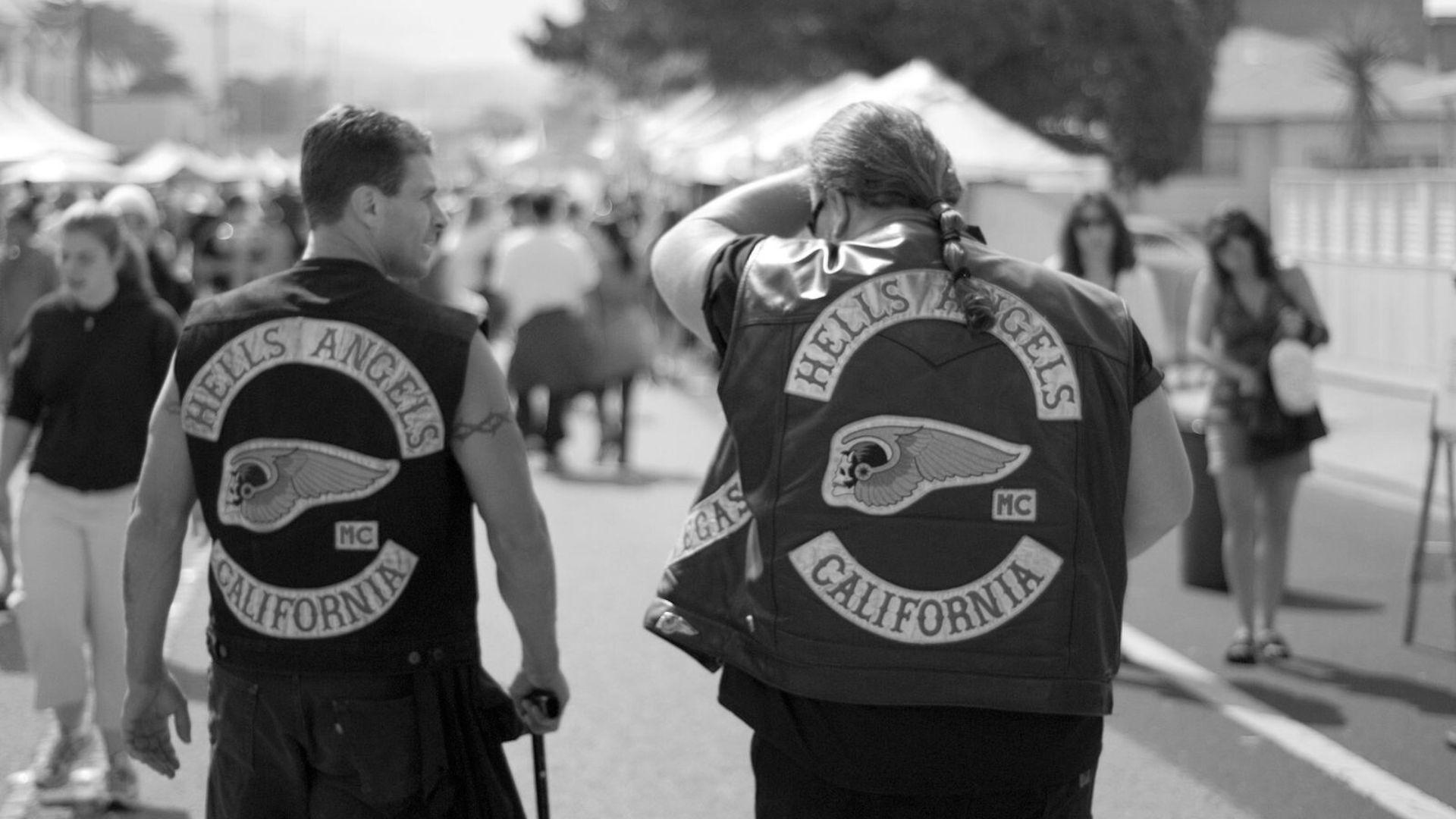 A black and white photograph showing two men walking away from the camera at an event. Both are wearing vests with large 'Hells Angels California' patches on the back. Around them, various other people are strolling
