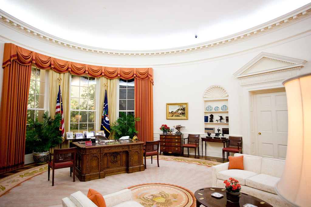 A white oval shaped office with orange curtains, couches, and a large dark wooden desk near the windows