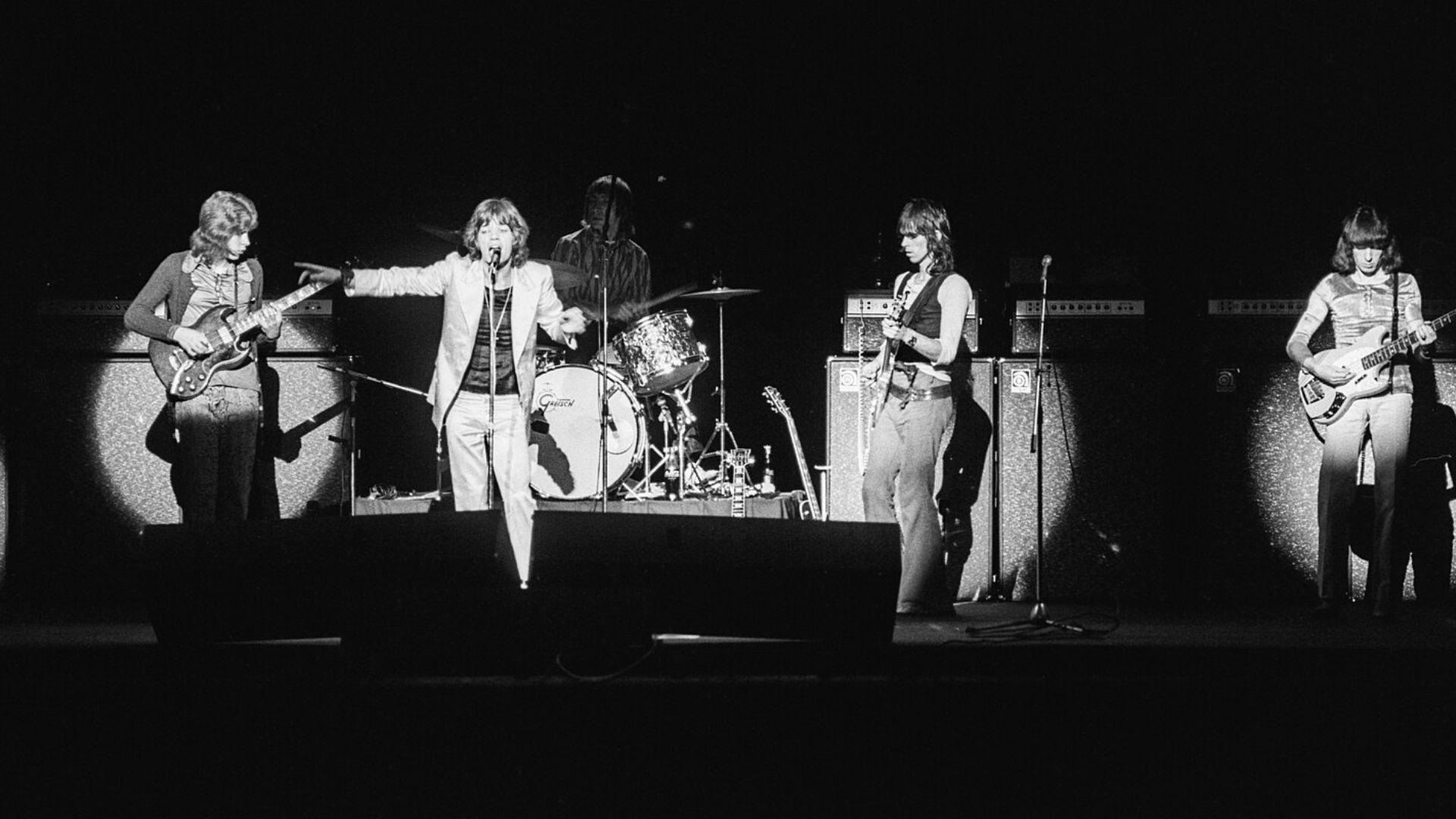 A black and white photograph of the Rolling Stones performing on a dark stage. The band consists of four members: a vocalist in the center with a hand outstretched, a drummer behind, and two guitarists on either side of the vocalist